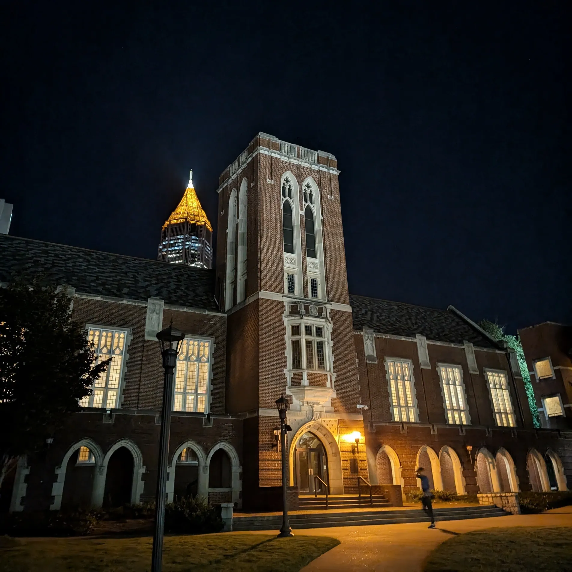 A gothic brick hall lit warmly at night, a gold-topped tower in the distance behind it.