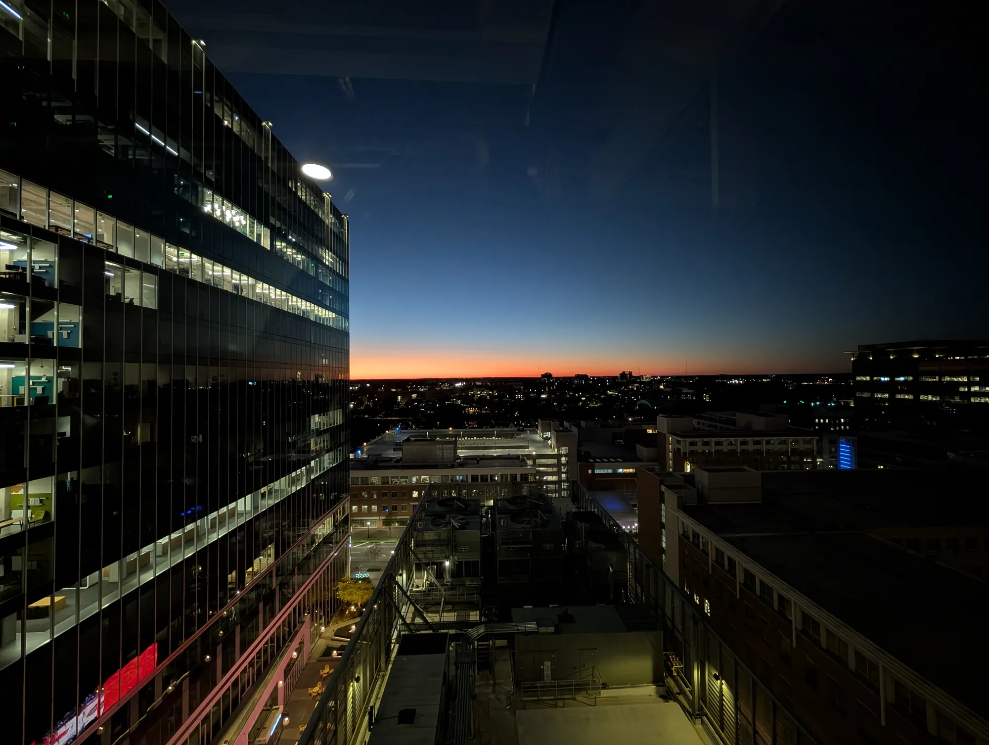 The city at dusk from a high window between glass buildings, a band of orange on the horizon.
