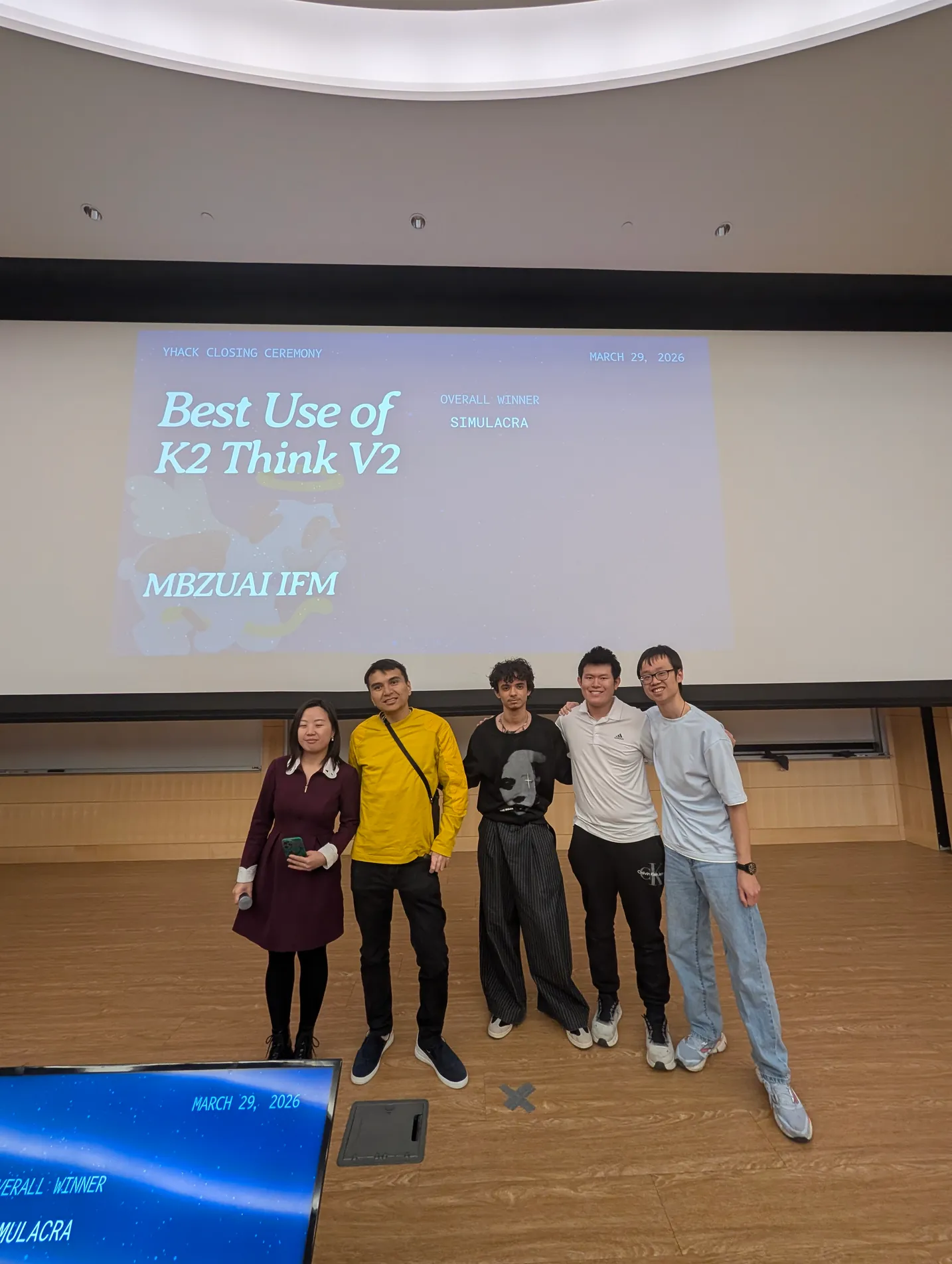 Five people in front of the YHack closing-ceremony slide for Best Use of K2 Think V2, naming Simulacra the overall winner.