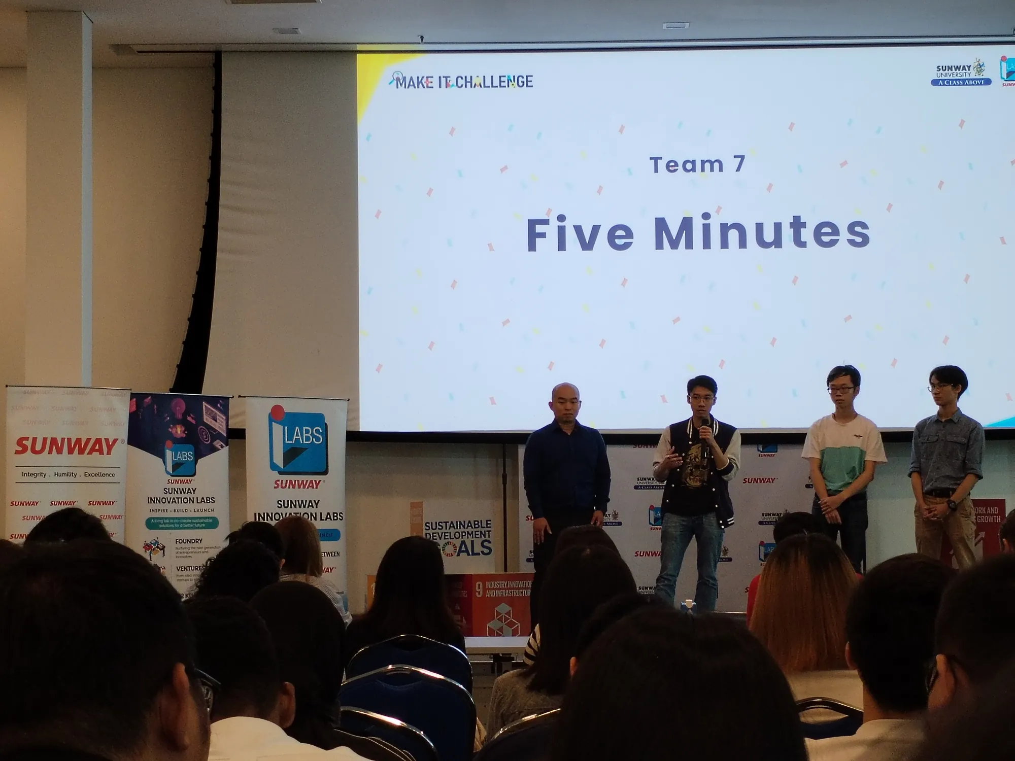 A team pitching on stage at the Make It Challenge, seen from the seated audience, under a large projected slide.