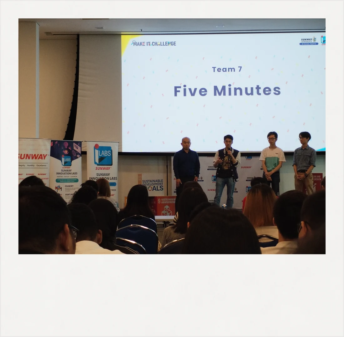 A team pitching on stage at the Make It Challenge, seen from the seated audience, under a large projected slide.